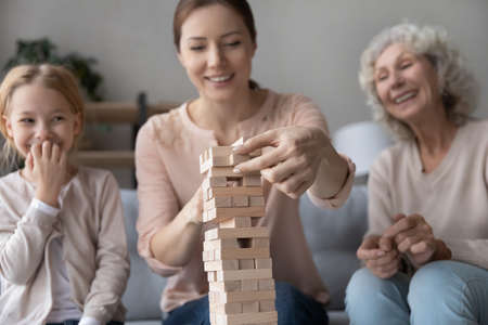 Close up three generations of women playing stack and crash board game together, smiling young woman with little daughter and mature mother building wooden tower, having fun on weekend at homeの写真素材