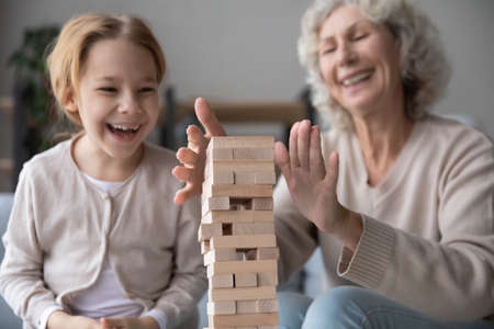 Close up laughing mature woman and little granddaughter building tower from wooden blocks, happy elderly grandmother and grandchild playing stack and crash board game, enjoying leisure time togetherの写真素材