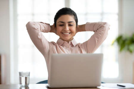 Smiling young indian female employee relax at workplace watch webinar or funny video on laptop. Happy millennial ethnic woman rest enjoy break at desk in office look at computer screen finish work.の写真素材