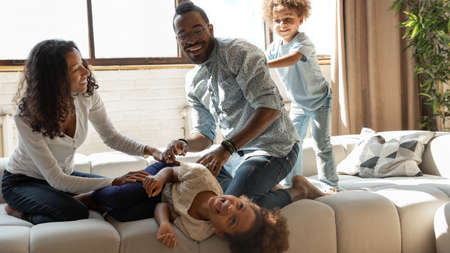 Overjoyed african american family involved in funny domestic activity on comfortable couch at home, happy mixed race father and mother tickling cheerful small daughter, having fun with kids.の写真素材