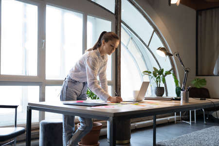 Concentrated young caucasian female executive preparing documents for project presentation alone in office, writing ideas or reminder or memo stickers, using creative approach in workflow management.の写真素材