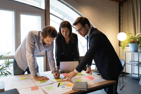 Focused male team leader leaning over table with multiracial colleagues, managing working processes together at briefing meeting, setting reminders on sticky paper notes, analyzing marketing reports.の写真素材