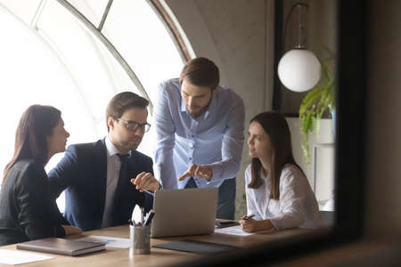 Focused young mixed race colleagues showing project presentation or marketing research results on computer to serious thoughtful 30s male team leader boss in formal wear, working together at table.の写真素材