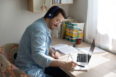 Happy young smart guy in glasses and wireless earphones looking at copybook notes, listening to educational lecture, enjoying study on online courses remotely at home office, e-learning concept.の写真素材
