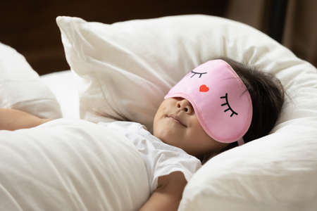 Close up cute Asian little girl wearing pink mask sleeping, taking nap in cozy bed alone, tranquil pretty toddler child resting on soft pillow under warm blanket duvet in bedroom at homeの写真素材
