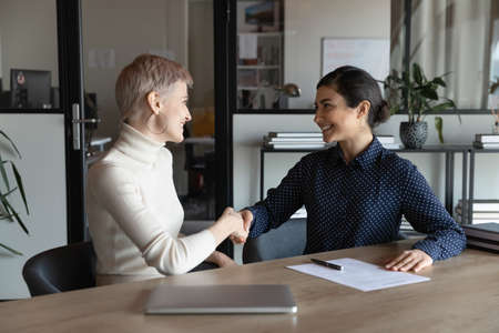 Happy multiracial female colleagues shake hands get acquainted greeting at business meeting in office. Smiling diverse businesswomen handshake close deal or make agreement. Partnership concept.の写真素材