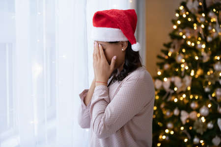 Unhappy African American woman in red Santa hat stand near window feel depressed lonely on Christmas Eve at home. Upset biracial female loner outcast celebrate New Year alone, depressed on holiday.の写真素材