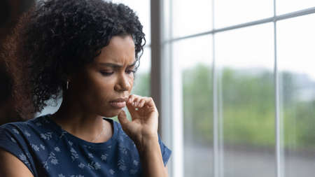 Close up pf unhappy 20s biracial woman stand near window miss or mourn at home. Sad depressed young...