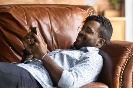 Happy young African American man lying relaxing on couch at home texting messaging online on cellphone. Smiling 20s biracial guy rest on sofa in living room browse wireless internet on smartphone.の写真素材