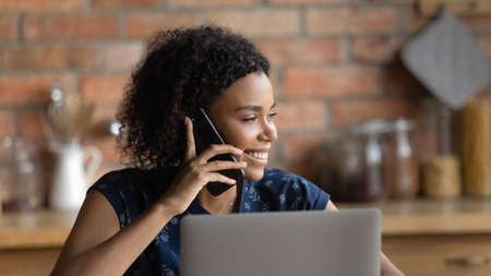Smiling young biracial woman talk have call on smartphone work on laptop look in distance thinking. Happy African American customer client use modern gadgets cellphone and computer at home office.の写真素材