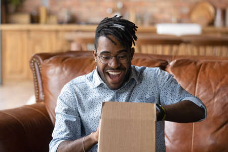 Overjoyed young African American man unpack cardboard box triumph with good quality order. Happy 20s biracial male unbox carton package with internet delivery, shopping on web online from home.の写真素材