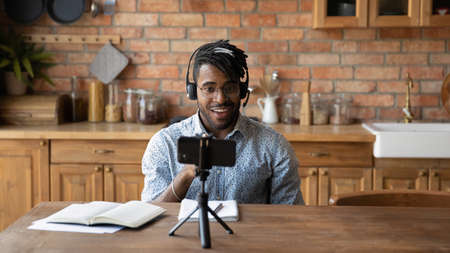 Young African American male vlogger influencer in headphones record video blog on smartphone at home. Millennial biracial man blogger talk speak shoot live broadcast on cellphone for social media.の写真素材