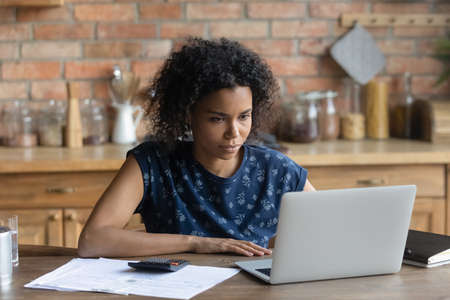 Focused young African American woman sit at desk at home look at laptop screen manage household...