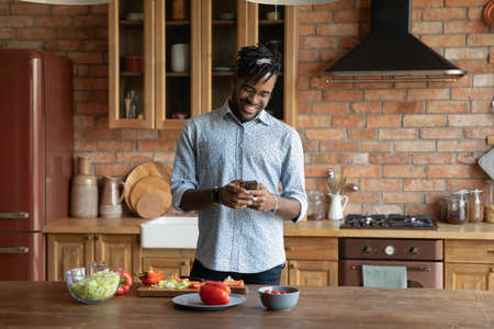 Smiling young African American man texting messaging on cellphone cooking in the morning at home kitchen. Happy 20s biracial male tenant renter use smartphone prepare salad for breakfast or lunch.の写真素材