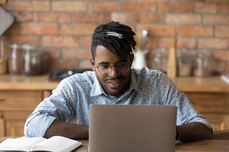 Millennial African American male student in glasses look at laptop screen study online on laptop at home. Young biracial man work distant at home office use computer, take web course or training.の写真素材