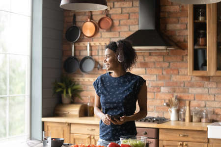 Happy young African American woman in headphones listen to music on cellphone cooking at home. Smiling 20s biracial female enjoy good sound in earphones and smartphone, prepare food in kitchen.の写真素材