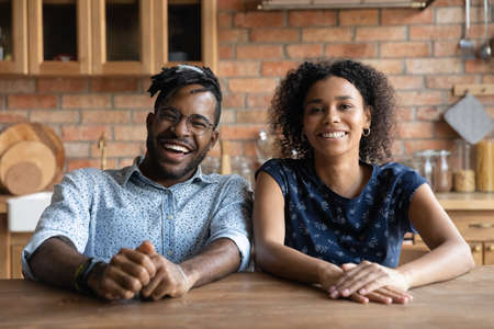 Portrait of overjoyed African American couple sit at home kitchen talk on video call together. Smiling biracial man and woman have fun laugh speak on webcam conference. Communication concept.の写真素材