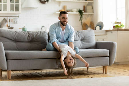 Loving young father playing with daughter at home, happy dad holding cute little girl upside down, sitting on cozy couch in living room, family playing funny game, spending leisure time togetherの写真素材