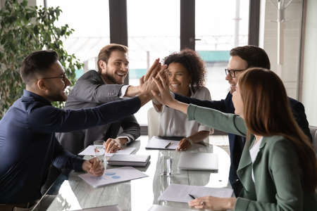 Overjoyed young mixed race business people sitting at table, joining hands in air, showing group unity, celebrating successful teamwork or professional achievement, raising team spirit at meeting.の写真素材