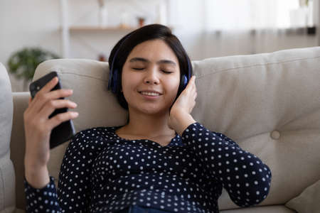 Calm happy young Vietnamese woman in wireless earphones relax on sofa at home enjoy good quality...