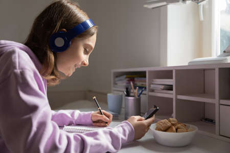 Happy smart Caucasian teenage girl in headphones make note handwrite study distant form home using smartphone. Smiling teenager in earphones look at cellphone screen write prepare homework online.の写真素材
