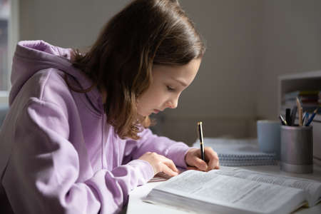 Concentrated Caucasian teen girl pupil handwrite in notebook prepare homework assignment at home. Focused smart teenager write read study with textbook at home. Distant education concept.の写真素材