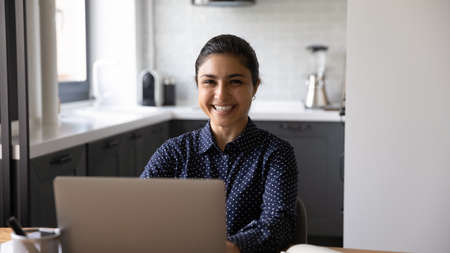 Portrait of smiling young Indian woman sit at desk in home office work on laptop online. Happy mixed...