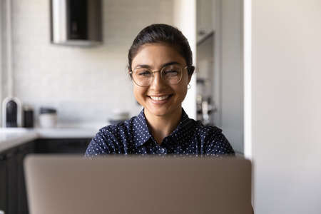 Smiling millennial Indian female in glasses look at laptop screen working online at home office. Happy young ethnic woman in spectacles typing on computer, consult client or study distant on web.の写真素材