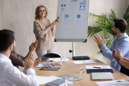 Excited diverse employees clap hands thank smiling middle-aged female speaker or coach for good...