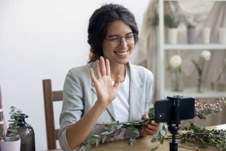 Happy young Caucasian woman florist wave greeting shoot video tutorial on smartphone. Smiling female...