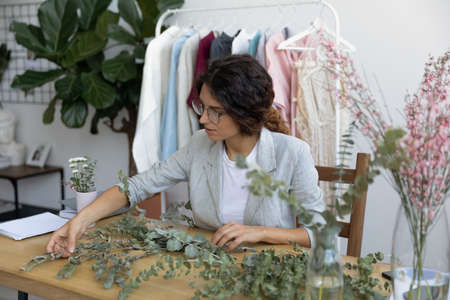 Focused millennial female designer make floral compositions or decorations on desk in cozy home...