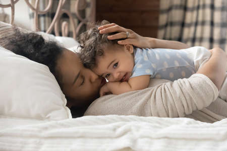 Loving young biracial mom lying in cozy bed hug cuddle cute little baby toddler. Caring happy...
