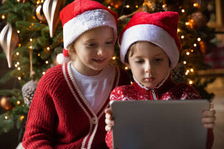 Close up of cute little boy and girl children in red Santa hats have fun watch video on tablet gadget at home. Smiling small brother and sister kids enjoy winter holidays play on new pad gift present.の写真素材