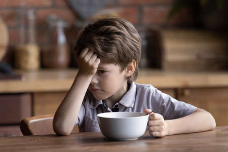 Unhappy small Caucasian boy child sit at kitchen table have no appetite for healthy breakfast. Sad...