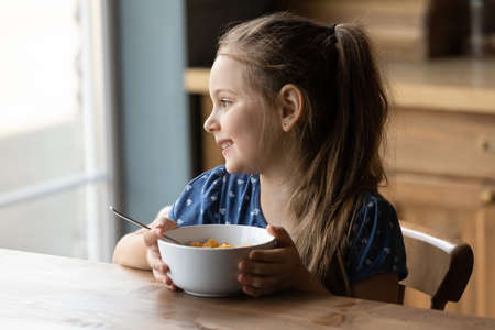Happy little 6s girl sit at table at home kitchen eat healthy cereals with milk look in distance dreaming or thinking. Smiling small kid child have delicious tasty nutritious breakfast. Diet concept.の写真素材