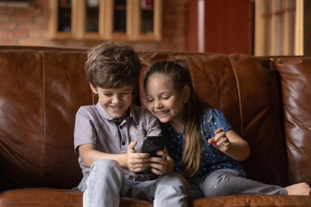 Happy cute little boy and girl children sit rest on couch at home look at cellphone screen play online game on gadget. Smiling small brother and sister kids have fun talk on video call on smartphone.の写真素材