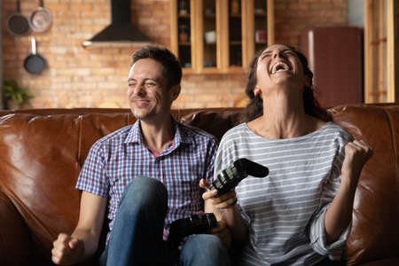 Overjoyed young Caucasian couple have fun engaged in funny gadget entertainment app at home. Excited millennial man and woman laugh celebrate win playing video computer game on device.の写真素材