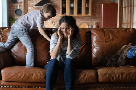 Unhappy young Caucasian mom suffer from headache stressed tired by naughty small 7s son playing. Upset exhausted mother feel distressed by ill-behaved naughty child kid, struggle from migraine.の写真素材