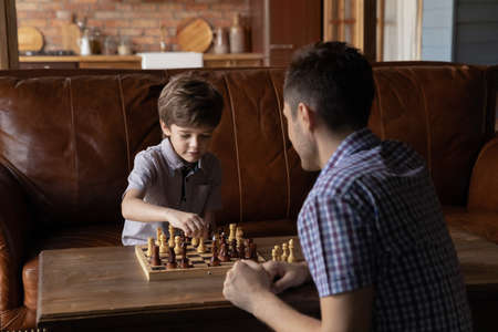 Small smart Caucasian boy child have fun playing chess with caring young dad at home. Focused little kid engaged in board game with father, think ponder over strategy. Children development concept.の写真素材
