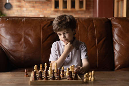 Smart little 7s Caucasian boy sit on couch at home play chess thinking pondering. Thoughtful pensive small kid engaged in board game, make decision, develop strategy. Children development concept.の写真素材