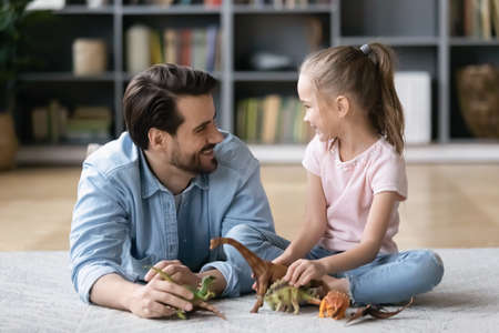 Smiling caring young father lying on floor carpet, enjoying playing toys with cute energetic little 6 years old kid daughter in living room, happy two generations family entertaining together at home.の写真素材
