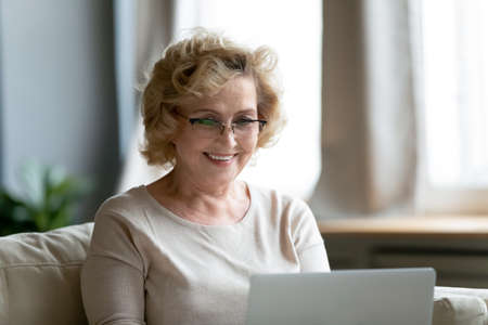Pleased happy middle aged mature woman in eyeglasses looking at computer screen, reading good media news, enjoying communicating in social networks with friends or grown children online at home.の写真素材