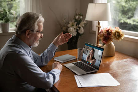 Mature man wearing glasses making video call to adult daughter, talking, chatting online, senior teacher mentor teaching student, explaining, working at home, using laptop and social media appの写真素材