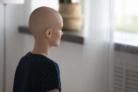 Close up rear view pensive hairless woman struggling with oncology, looking in window, sitting alone, thoughtful upset young bald female patient suffering from cancer, healthcare and treatmentの写真素材