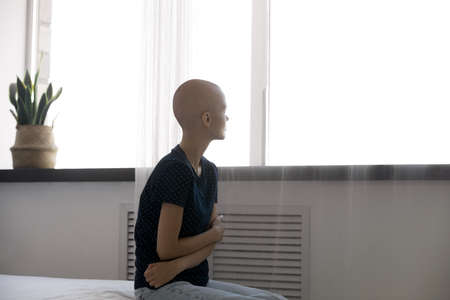 Rear view hopeless hairless woman struggling with cancer, oncology, sitting on bed alone, hugging herself, looking to aside, thinking about health problem, dreaming about recovery, remissionの写真素材