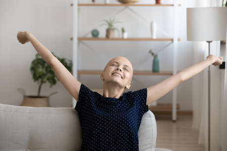 Close up smiling hairless woman stretching hands on couch, relaxing, enjoying lazy leisure time, happy young female with closed eyes and hands outstretched sitting on sofa.の写真素材