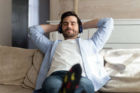 Peaceful weekend. Happy carefree young millennial man resting on sofa at home lying in relaxed pose alone putting headphones.の写真素材