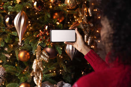 Rear view African American woman holding phone with empty white screen mockup on Christmas tree...
