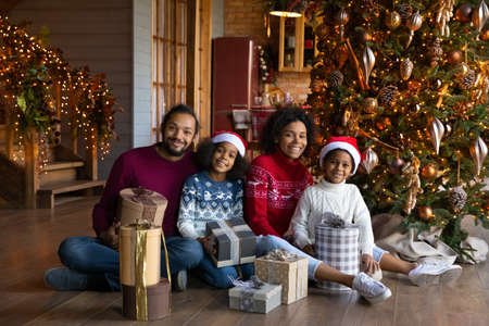 Portrait happy African American family wearing festive red caps and warm sweaters celebrating Christmas, smiling parents with daughter and son sitting on warm floor with gift boxes, looking at cameraの写真素材