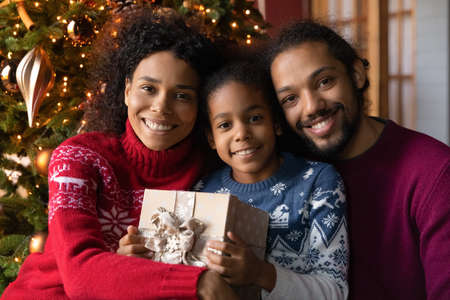 Head shot portrait happy African American family celebrating Christmas, sitting near festive tree, smiling mother and father hugging adorable little daughter holding gift box, looking at cameraの写真素材